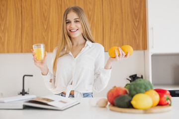 A happy blonde woman is looking at a recipe book and drinking juice in the kitchen