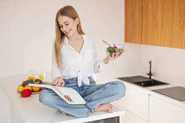 Happy and smiling blonde woman sitting on top of table in kitchen and reading recipe book and...