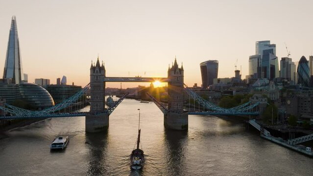 The Sun Is Setting Over The City Of London As A Sail Boat Passes Under Tower Bridge
