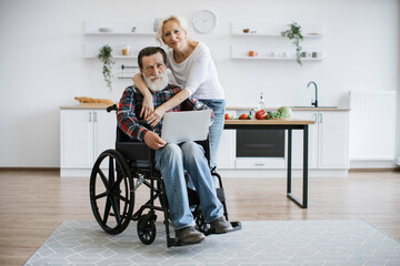 Charming old couple of man in wheelchair with laptop and wife hugging him on background of bright modern kitchen, looking at camera. - obrazy, fototapety, plakaty