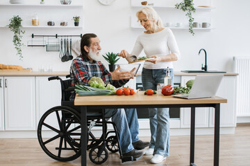 Senior couple, man in wheelchair and elderly woman, making healthy salad. Male with gray beard...