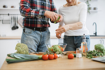 Close up of hands of elderly man and woman in casual clothes tears lettuce and herbs into glass bowl. Old retired couple preparing fresh healthy salad in modern kitchen at home.