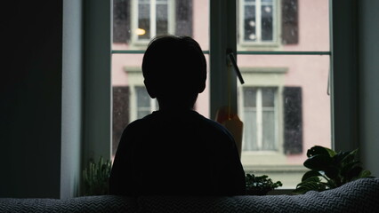 Silhouette of child staring at snow fall from home window during winter season. Back of young boy...