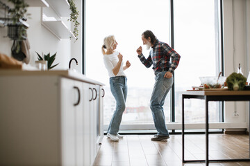 Caucasian old couple spends free time listening to music and dancing the twist in modern light kitchen. Seniors husband and wife having fun at the weekend in a cozy house. - obrazy, fototapety, plakaty