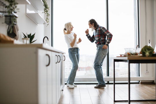 Caucasian old couple spends free time listening to music and dancing the twist in modern light kitchen. Seniors husband and wife having fun at the weekend in a cozy house.
