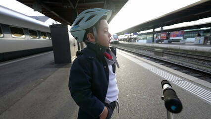 Obraz premium One pensive young boy standing at train platform wearing helmet staring blankly into the horizon daydreaming. Thoughtful Child with hands in pocket waiting for transportation to arrive