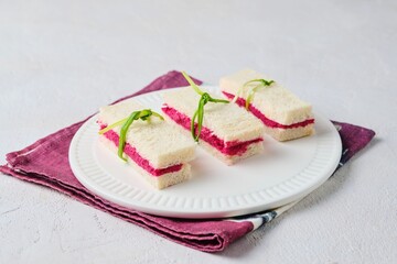 Tea sandwiches on white bread with beetroot hummus on a light ceramic plate on a light concrete background. Sandwiches for tea