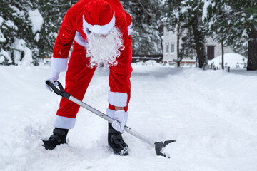Naklejka premium Santa Claus cleans snow with shovel in winter outdoors after a snowfall. Cleaning the streets in the village, clearing the passage for cars, difficult weather conditions for Christmas and New Year