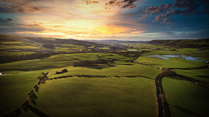 Aerial view of a lush green countryside at sunset with rolling hills, fields, and a winding river.