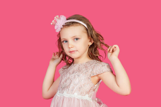 Portrait of positive little girl posing gesture hands at pink background, looking at camera. Perfect kid lady with blue eyes in pink dress, studio shot. Child image emotion concept. Copy ad text space