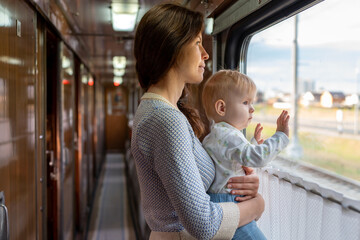Mom holding her infant baby girl in her arms while riding in a public transportation. Cute toddler girl traveling with his mother. Family traveling in a train and looking through window. - obrazy, fototapety, plakaty