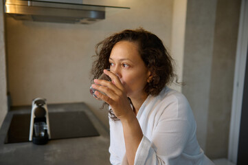 Close-up portrait of a Latin American young adult confident pretty woman in white robe, drinking water in the morning.