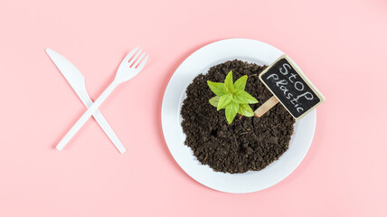 Plastic knife and fork crossed, white plate with black earth, green sprout and peg with the inscription: stop plastic.