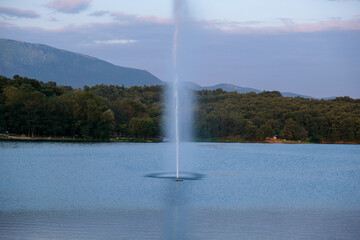 Tirana lake with a waterjet and breeze from a fountain, hills and green leafy trees on misty background.