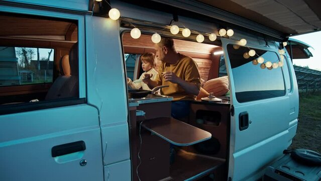 Under the string lights, a man and a child engaged in cooking inside a camper van, solar plant at the backgrount in the evening dusk