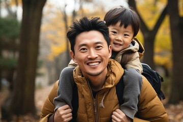 Happy Asian father carrying his son on his back in the park - obrazy, fototapety, plakaty