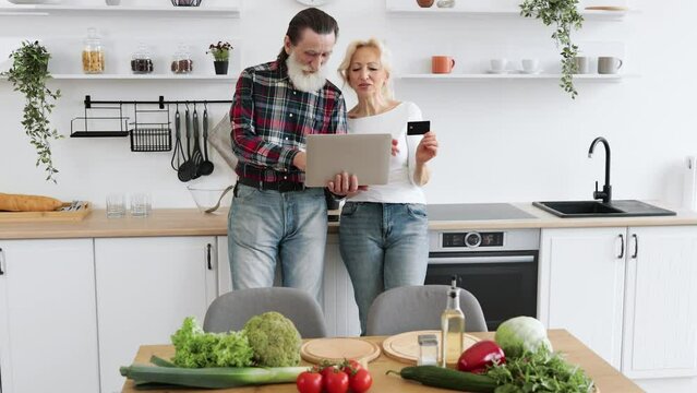 Old Caucasian couple using laptop and credit card to buy food products to prepare breakfast in modern spacious kitchen background.