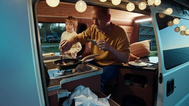Inside the cozy camper van, a boy and his grandfather bond over the simple joy of cooking and eating together at the dusk. Solar power pland at the background