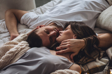 Obraz na płótnie Canvas Beautiful happy loving young smiling couple relaxing in bed, looking at each other. Cozy home atmosphere, tenderness, closeness. Embracing, kissing. Lazy weekend, slow living concept