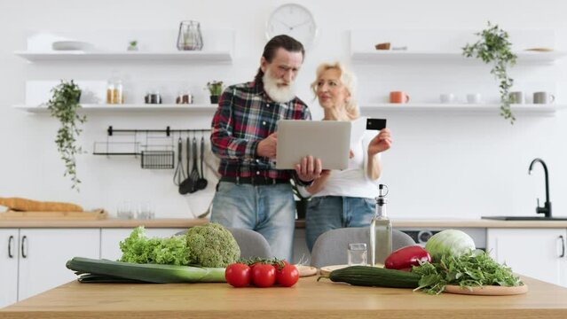 Focus on the vegetables and herbs lying on the table. Old Caucasian couple using laptop and credit card to buy food products to prepare breakfast in modern spacious kitchen background.