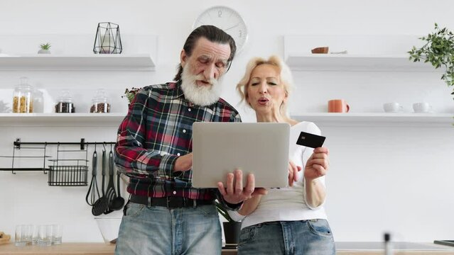 Focus on laptop in hands of happy elderly husband and wife holding credit card paying for food to prepare delicious breakfast in modern light kitchen background.