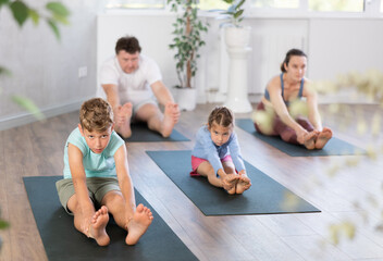 Diligent preteen boy sitting on the floor and practicing stretching on black mat together with his parents and sister