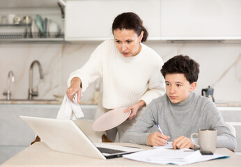 Teen son doing school homework while mom washes dishes in the kitchen