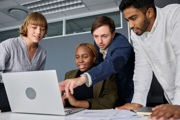 Four young adults in businesswear talking and gesturing while working on laptop at desk in office