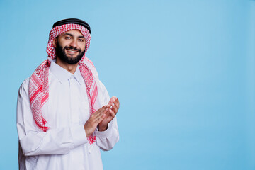 Cheerful smiling muslim person wearing traditional clothes clapping hands and looking at camera. Excited arab man showing applause gesture studio portrait on blue background