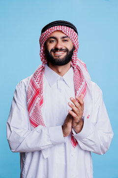 Excited muslim man in thobe and ghutra standing confidently, applauding with hands studio portrait. Smiling arab person clapping arms at performance and looking at camera