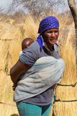 African woman carrying the baby in the back wrapped in a towel the traditional way, thatch grass in the background , village life.