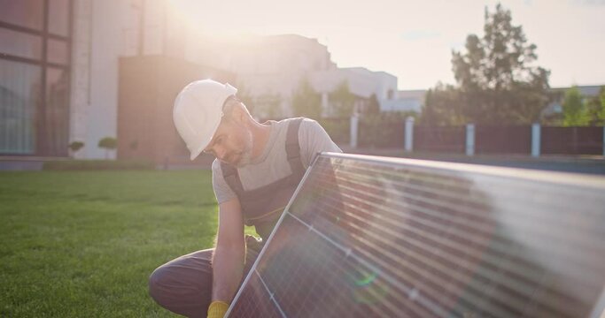 Caucasian man working at renewable energy enterprise. Professional contractor crouching at lawn. Male technician engineer installing solar panels. Conversion of sunlight into electricity.