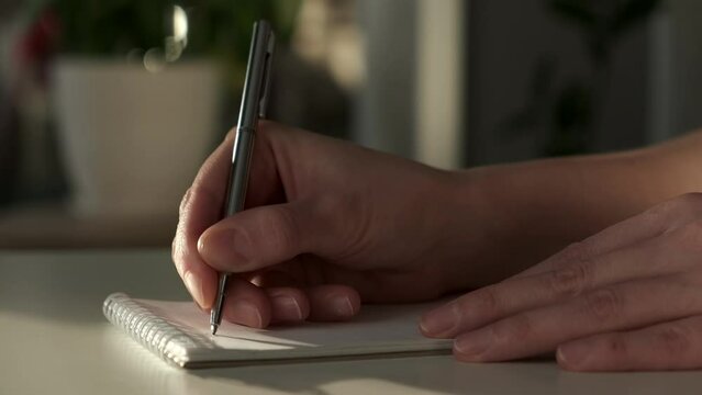 Closeup of woman's hands journaling in a white notebook on a white table at home.