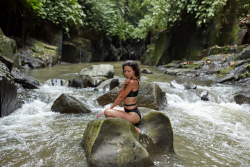 A young slender woman in a swimsuit poses on a huge boulder covered with moss in the middle of a mountain river in the jungle on the popular island of Bali.