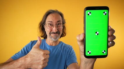 Excited mature elderly man standing and pointing Big smartphone with blank green screen with...