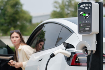 Young woman use smartphone to pay for electricity at public EV car charging station green city...