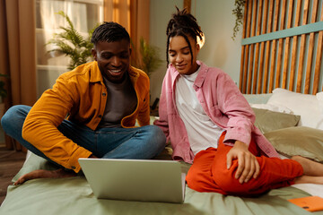 Smiling attractive couple African American young man and woman sitting on bed at home