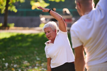 A group of seniors follows a trainer, engaging in outdoor exercises in the park, as they...