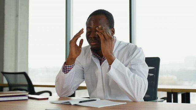 Stressed frustrated black doctor in white uniform with headache sitting alone at work desk in hospital. Sad young gp feeling desperate and guilty for medical failure
