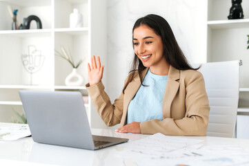 Positive successful indian or arabian business lady, company employee sitting at workplace in the office, using laptop for online video conference with business team or clients, greeting, smiling