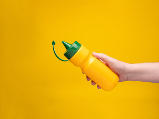 A squeeze bottle for mustard or sauces is held in the hand. Yellow background, no face. 