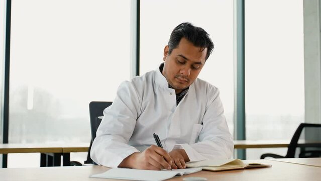 Stressed frustrated Indian doctor with headache sitting alone at work desk in hospital. Sad young gp feeling desperate and guilty for medical failure