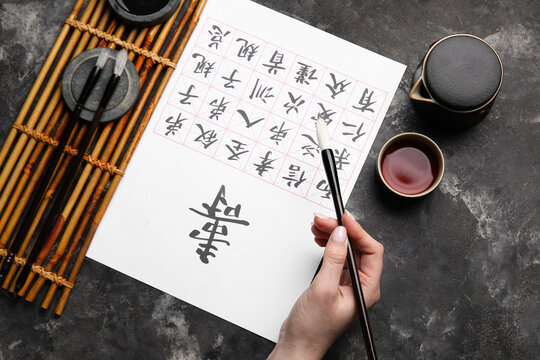 Female hand with Asian hieroglyphs, brushes and cup of tea on grunge background, closeup. International Haiku Poetry Day