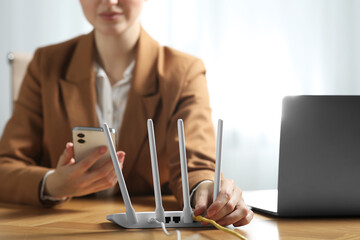 Woman with smartphone connecting cable to Wi-Fi router at table indoors, closeup