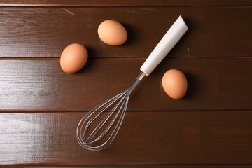 Metal whisk and raw eggs on wooden table, flat lay