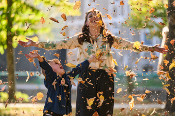 A modern woman joyfully plays with her son in the park, tossing leaves on a beautiful autumn day,...