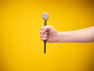 A  hand holds a flat chisel for stone work. No face, yellow background. 
