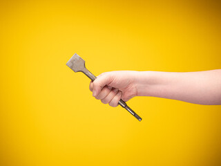 A construction worker's hand holds a flat chisel for stone work. No face, yellow background. 
