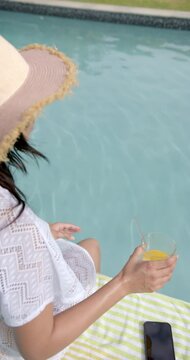 Vertical video of biracial teenage girl in sunhat sitting by sunny pool with juice, slow motion