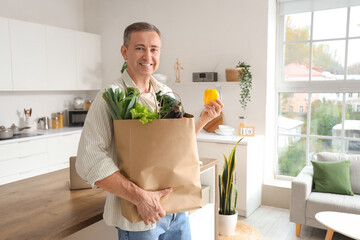 Mature man with paper bag of fresh vegetables in kitchen - obrazy, fototapety, plakaty